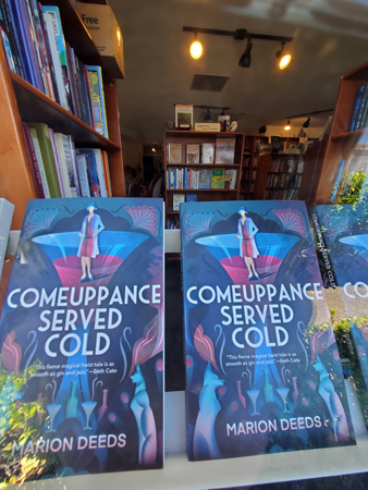 View through window to back of store. Two copies of Comeuppance Served Cold in foreground