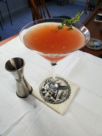 Pink drink in martini glass with thyme spring garnish, jigger to left